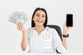 Photo of lucky businesswoman 30s holding smartphone and fan with dollar money banknotes while sitting in armchair in office