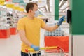Photo of male consumer uses touchscreen in shop, checks price, poses with shopping cart, wears disposable mask and gloves, pays