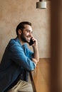 Photo of masculine satisfied man smiling and talking on cellphone while sitting on chair in cafe indoors Royalty Free Stock Photo