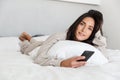 Photo of mature woman 30s using mobile phone, while lying in bed with white linen in bright room Royalty Free Stock Photo