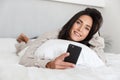 Photo of middle-aged woman 30s using mobile phone, while lying in bed with white linen in bright room Royalty Free Stock Photo