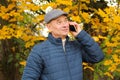 Photo of an old man in a gray cap and a blue jacket, who is standing in the autumn park, is phoning and smiling, the concept of Royalty Free Stock Photo