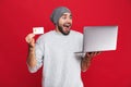 Photo of optimistic guy holding credit card and silver laptop isolated over red background