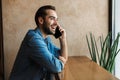 Photo of optimistic young man smiling and talking on cellphone while sitting on chair in cafe indoors Royalty Free Stock Photo