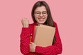 Photo of overjoyed Caucasian lady clenches fist, has broad smile, holds notepad, pencil, cheers as has success