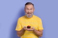 Photo of overjoyed funny pensioner man dressed striped t-shirt hands holding small homemade muffin isolated on purple Royalty Free Stock Photo