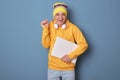 Photo of overjoyed smiling cheerful man wearing beanie hat and casual yellow hoodie, holding closed laptop, clenched fist,