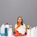 Photo of pensive Caucasian woman wears rubber protective gloves and headband, surrounded with bottles of detergents for laundry,