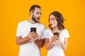 Photo of perplexed man and woman holding and peeking at cell phones, isolated over yellow background Royalty Free Stock Photo