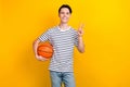 Photo of pleasant student dressed striped t-shirt hold bascketball ball showing v-sign symbol isolated on vivid yellow