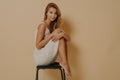 Photo of pleased young female model wears dress has slender legs crumpled up sits on chair