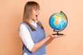 Photo portrait of amazed girl looking at globe in spectacles opened mouth isolated on pastel beige color background