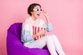 Photo portrait of amazed woman in 3d glasses with big popcorn bag sitting in violet beanbag chair isolated on pastel