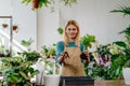Photo portrait blonde female owner in apron showing how to transplant a plant, holding soil, plant, smiling to camera at