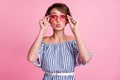 Photo portrait of pouting girl touching rose-tinted heart-shaped glasses on pastel pink colored background