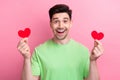 Photo portrait of young astonished man wearing green t shirt holding red paper postcards heart figures isolated on pink