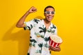 Photo of positive attractive guy wear stylish clothes hold basket tasty pop corn raise fist have fun isolated on yellow