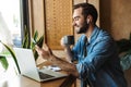 Photo of positive bearded man holding cup of tea and using smartphone while working in cafe indoors