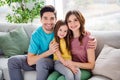 Photo of positive three people loving mother father hug embrace cuddle little girl kid sit sofa in house indoors