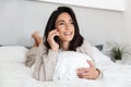 Photo of positive woman 30s using mobile phone, while lying in bed with white linen in bright room Royalty Free Stock Photo