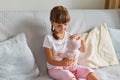 Photo of preschooler girl sitting on the sofa in the room at home playing with baby doll like mom, child wearing white t shirt and Royalty Free Stock Photo