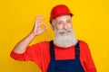 Photo of pretty cheerful senior guy dressed uniform overall red hardhat showing okey sign isolated yellow color Royalty Free Stock Photo