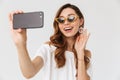 Photo of pretty european woman 20s wearing sunglasses taking selfie on smartphone, and touching earring, isolated over white back Royalty Free Stock Photo