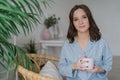 Photo of pretty female with appealing look, dressed in casual domestic clothes, holds white mug with aromatic coffee or tea, sits
