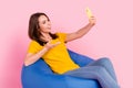 Photo of pretty sweet young lady wear yellow t-shirt sitting beanbag having video call modern gadget isolated pink color