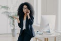 Photo of puzzled prosperous businesswoman calls partner, raises palm, holds mobile phone, wears spectacles and formal black outfit