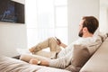 Photo of relaxed adult man 30s in casual clothing sitting on sofa in living room, and watching TV