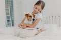 Photo of relaxed beautiful small girl plays with pedigree dog, embraces favourite animal, dressed in casual pyjamas, sits on bed,