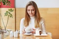Photo of relaxed light haired female drinks hot coffee or cappucino with delicious dessert, sits in cosy cafeteria, waits for