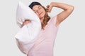 Photo of relaxed pleased attractive female stretches, holds pillow, wears sleep mask, has pleasant dreams, has slumber at afternoo Royalty Free Stock Photo