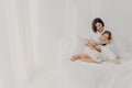 Photo of relaxed young mother embraces daughter with love, pose in pyjamas bare foot, have pleasant smiles, being in spacious