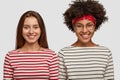 Photo of satisfied multiethnic women in striped sweaters, have pleasant smiles on faces, model against white background