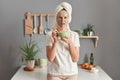 Photo of scared Caucasian woman with towel on head standing speechless indoor with beauty facial mask to reduce fine lines eats