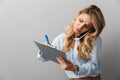 Photo of serious blond secretary woman with long curly hair writing down notes in clipboard while calling on smartphone