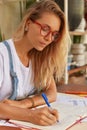 Photo of serious blonde woman journalist wears spectacles, writes down ideas in notepad, wears white t shirt and