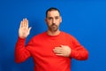 Photo of serious and confident bearded hispanic man with hand on chest promising oath isolated over blue colored