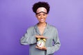 Photo shiny attractive african lady grey nightwear smiling holding arms showing clock isolated purple color background