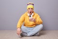 Photo of shocked amazed young adult man wearing beanie hat, yellow hoodie and jeans posing against gray wall, sitting on floor Royalty Free Stock Photo