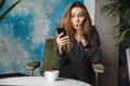 Shocked emotional beautiful young pretty woman sitting in cafe indoors using mobile phone chatting Royalty Free Stock Photo