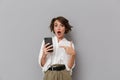 Photo of shocked woman 20s holding and using mobile phone, isolated over gray background