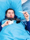 Photo of sick brunet man in scarf with thermometer in hand lying in bed.