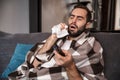 Photo of sick man 30s wrapped in blanket being ill while sitting on sofa in apartment