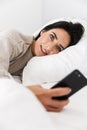 Photo of single woman 30s using smartphone, while lying in bed with white linen at home Royalty Free Stock Photo