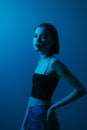 Photo of skinny attractive female model in blue light on wall background, posing at camera with serious face