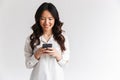 Photo of smiling asian woman with long dark hair holding and using black mobile phone, isolated over white background in studio Royalty Free Stock Photo