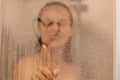 Photo of smiling attractive young woman draws heart on weeping glass shower door, enjoys rest in douche, washing her body,